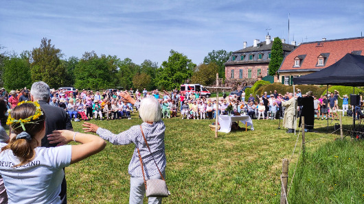 Gottesdienst im Garten von Schloss Wolfsgarten: Viele Menschen stehen auf der Wiese, die beiden Pfarrer unter einem Sonnenschutz, in der Mitte ein Kreuz aus zwei Birkenstämmen, im Vordergrund schenken Menschen ihre Arme im Takt zur Musik