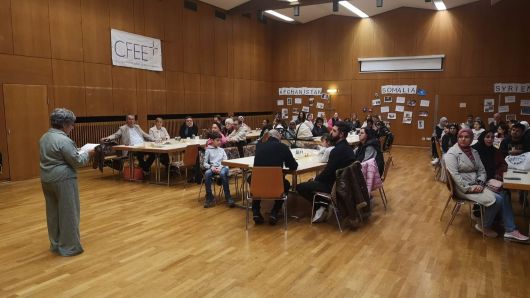 Teilnehmer an Tischen sitzend, werden von Frau Rezvani-Thürck begrüßt